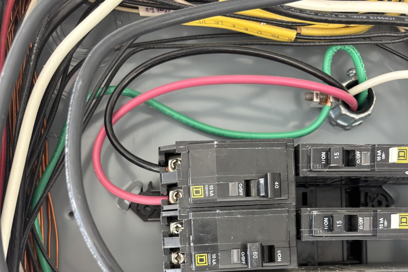 Inside a residential breaker panel: black, red and green conductors entering through a conduit fitting and landing on a two-pole breaker